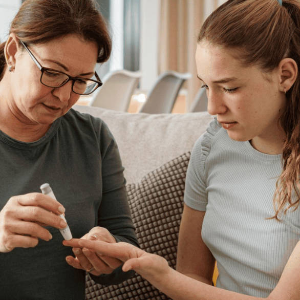 Een moeder prikt dochter met diabetespen in de vinger. De sfeer is huiselijk. 